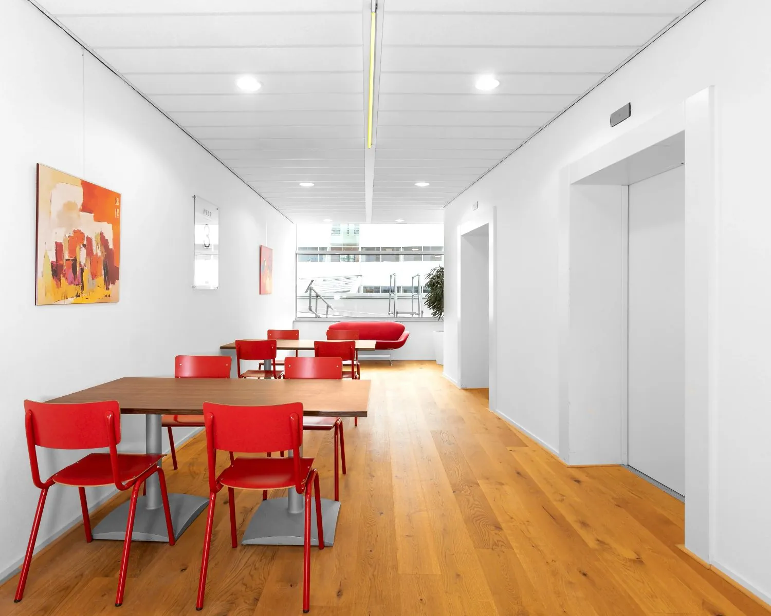 Moderne kantoorruimte met houten vloer, rode stoelen en tafels, en een lift aan de rechterzijde.