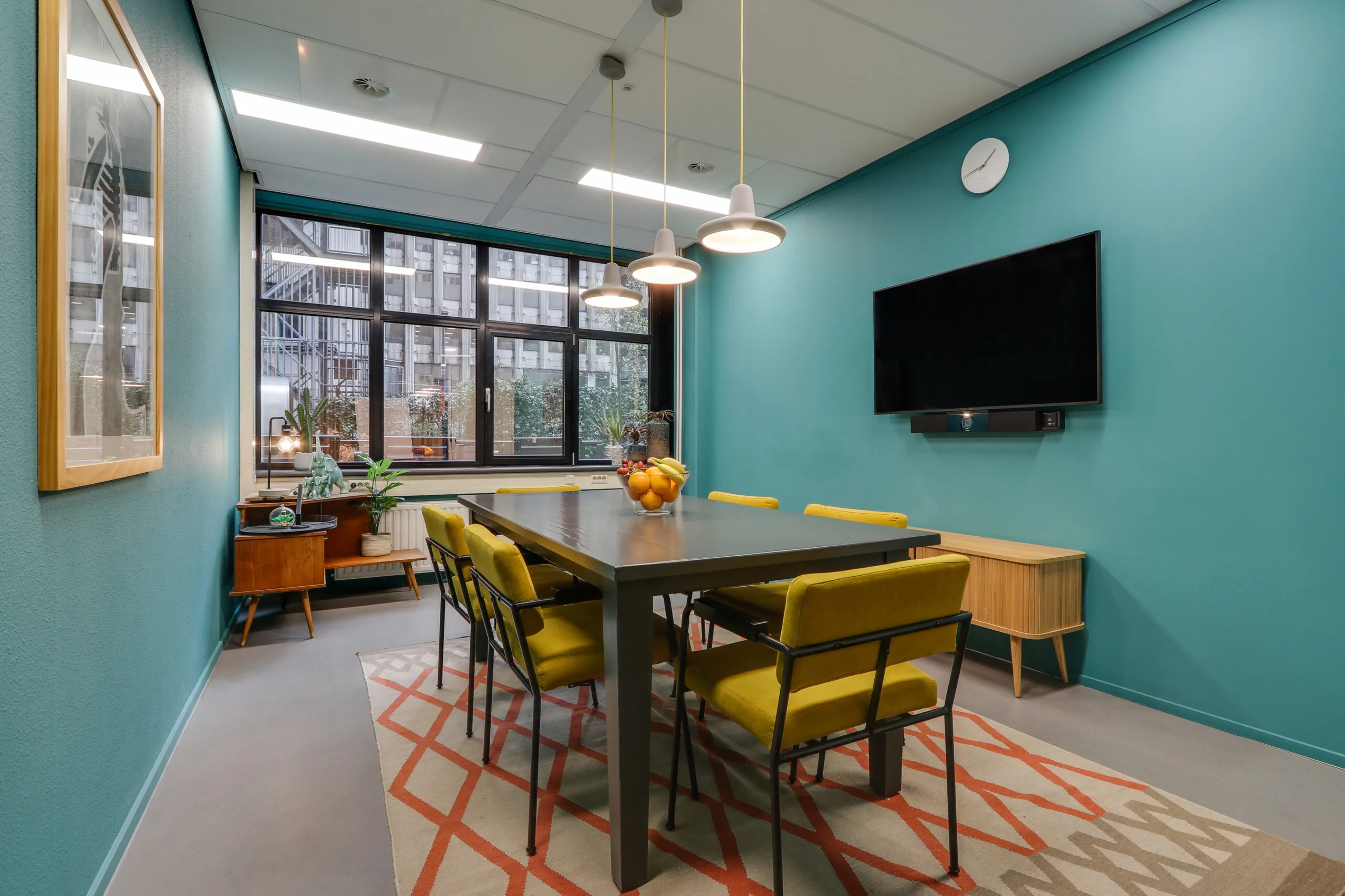 Vergaderruimte in Rotterdam Centrum met turquoise muren, gele stoelen, een zwarte tafel en een wandtelevisie.