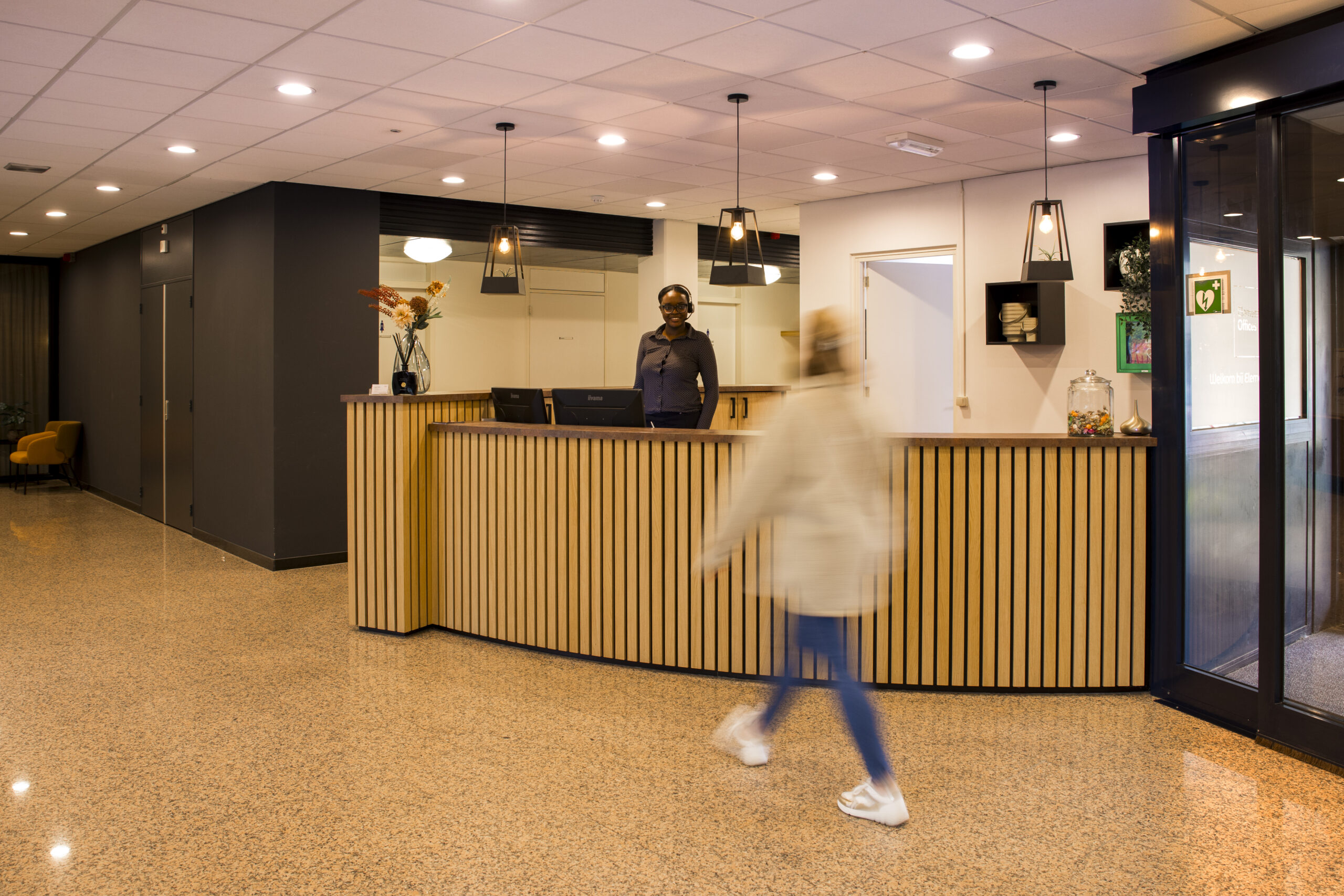 Receptie met een receptionist achter een houten balie en een persoon die voorbij loopt in een moderne entree.