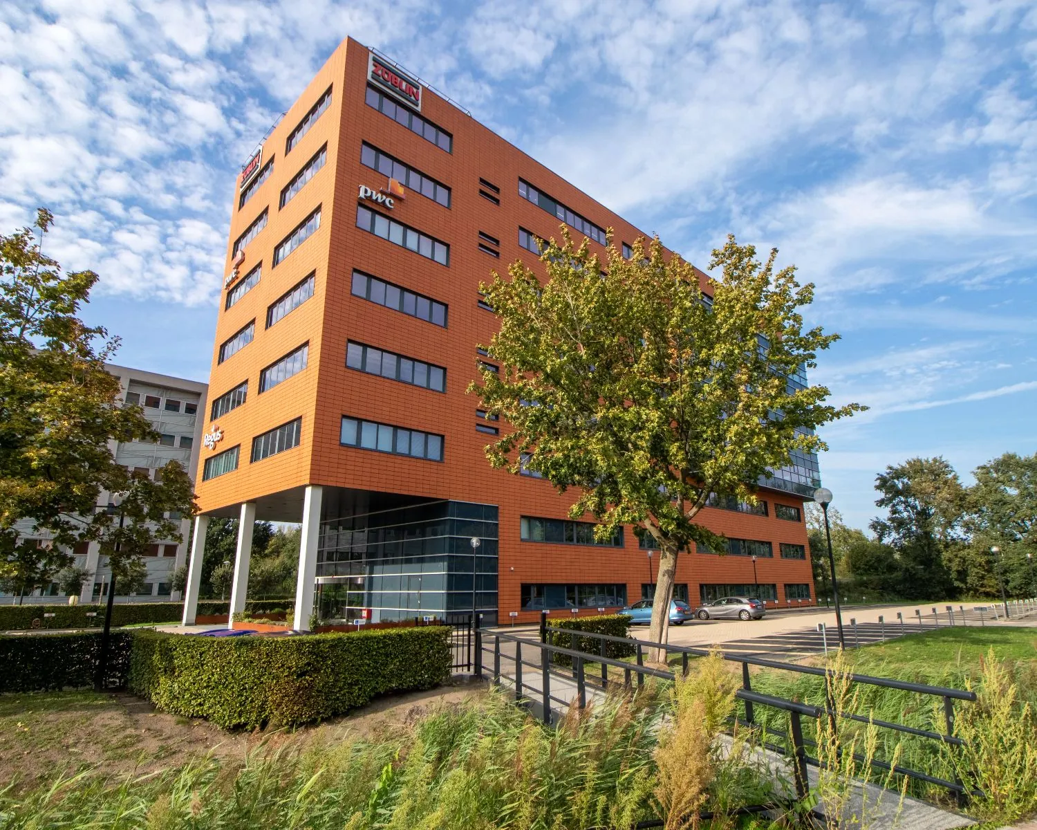 Modern kantoorgebouw 5018_1 met oranje gevel en logo's van onder andere PWC, omringd door groen en een parkeerplaats.