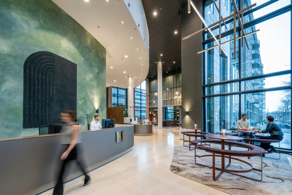 Een moderne hotellobby met een receptie, personen aan het werk en mensen in gesprek bij het raam.