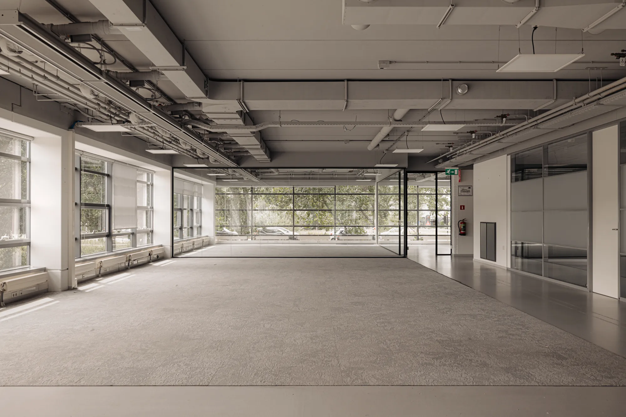 Een lege, moderne kantoorruimte met grote ramen en zichtbare leidingen aan het plafond.