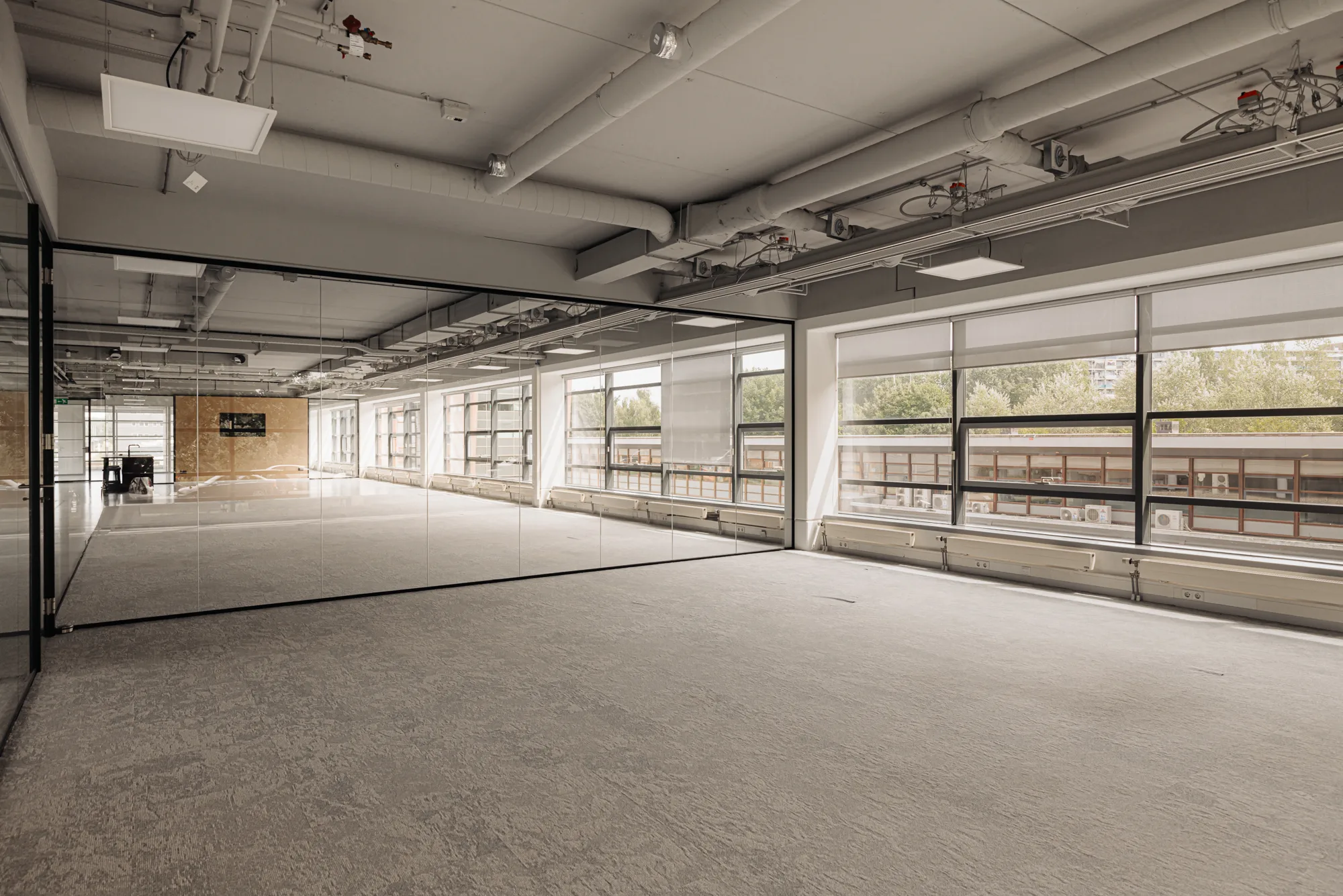 Een lege kantoorruimte met glazen wanden en veel ramen die uitzicht geven op bomen en een gebouw.