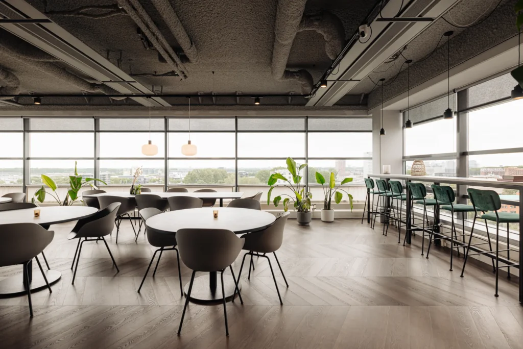 Een moderne, lichte kantoorruimte met ronde tafels, stoelen en hoge krukken bij grote ramen.