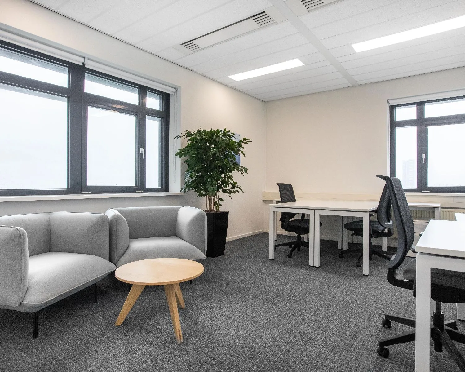 Modern ingerichte kantoorkamer met bureaus, zithoek en grote ramen.