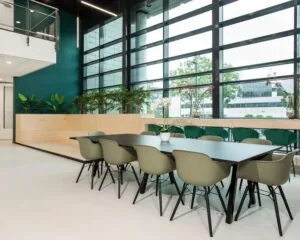 Moderne kantoorpresentatieruimte met een lange vergadertafel, groene stoelen en grote ramen met uitzicht op een bedrijfsgebouw.