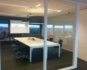 Vergaderruimte met grote tafel, bureaustoelen en zicht op een kantoorruimte met ramen.