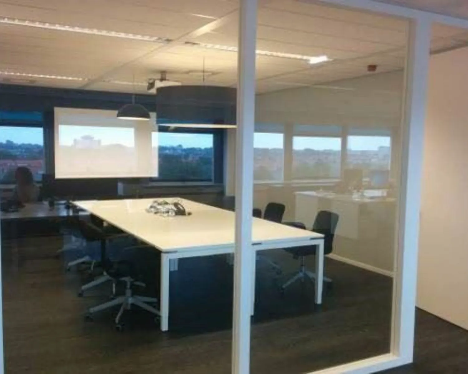 Vergaderruimte met grote tafel, bureaustoelen en zicht op een kantoorruimte met ramen.