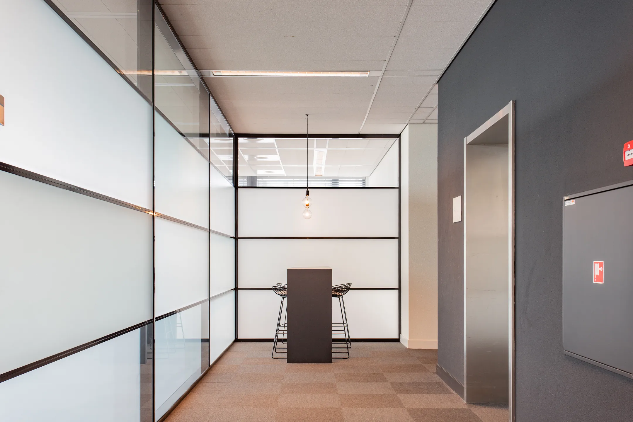 Een moderne kantoorruimte met een hoge tafel en krukken, omgeven door glazen panelen en een hangende lamp.