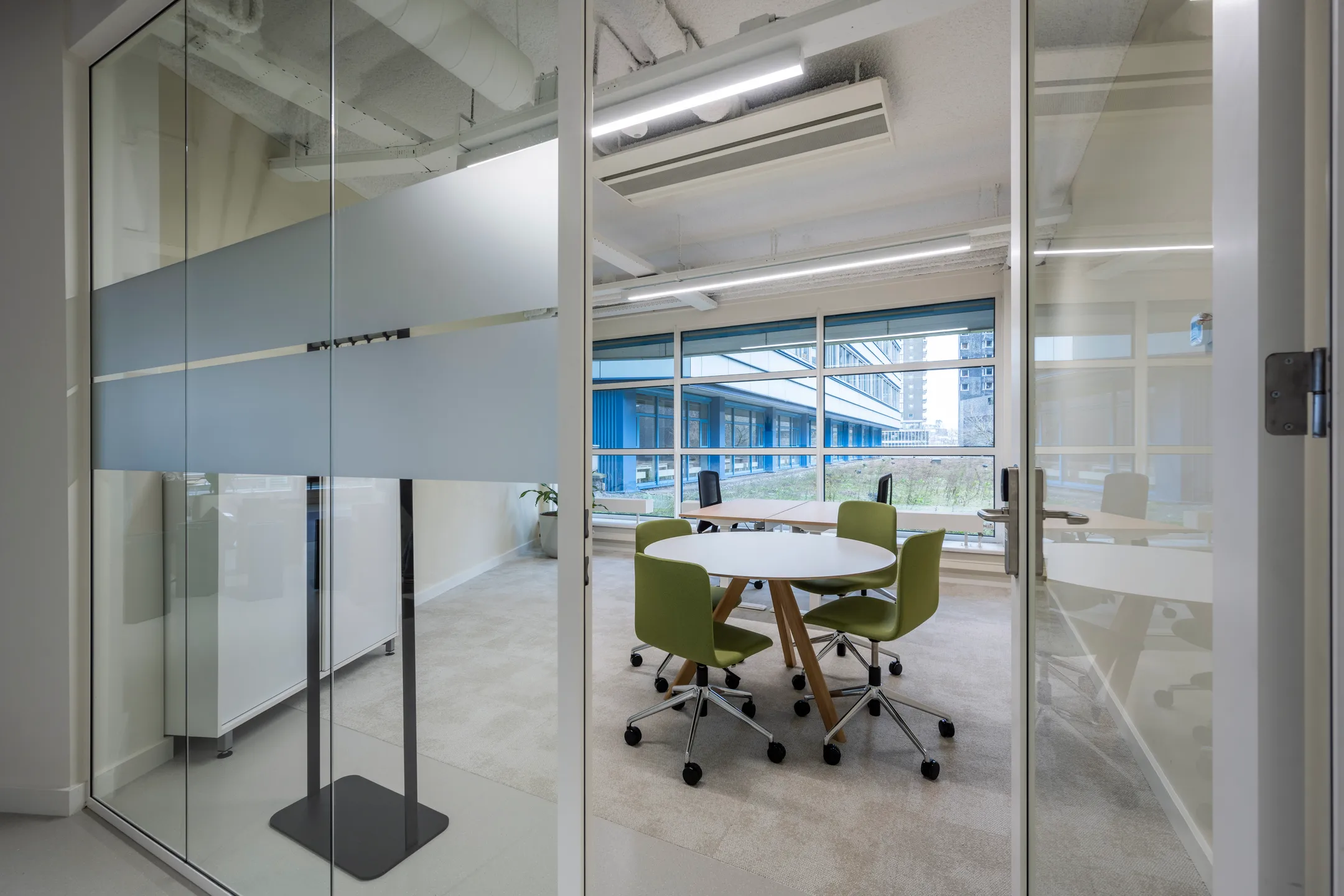 Moderne vergaderruimte met groene stoelen en een ronde tafel, gezien door een glazen wand.