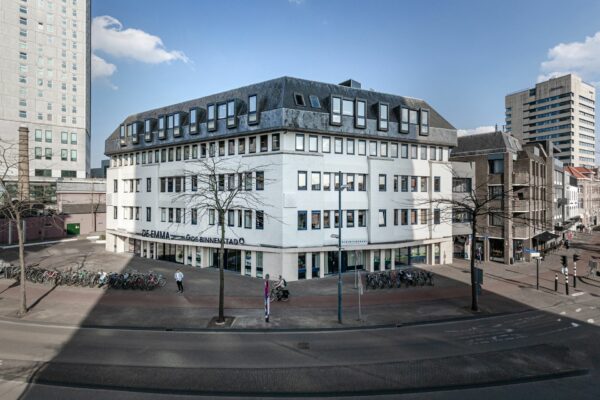 Wit kantoorgebouw De Emma aan een drukke straat in de binnenstad met fietsenrekken en enkele voorbijgangers.