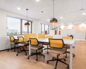 Modern kantoorinterieur met gedeelde werkplekken, gele bureaustoelen en hanglampen in een lichte ruimte.