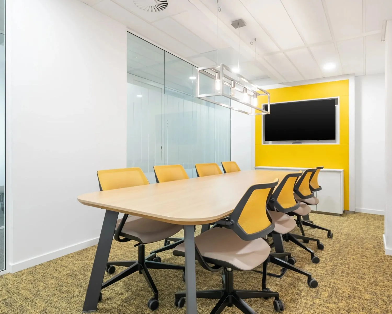 Vergaderruimte met een houten tafel, zes gele stoelen, een grote monitor aan de muur en moderne verlichting.