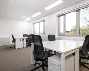 Moderne, lichte kantoorruimte met witte bureaus en zwarte bureaustoelen bij grote ramen.