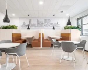 Modern ingerichte kantoorlounge met zitbanken, ronde tafels, grijze stoelen en een wandillustratie van een stadsgezicht.