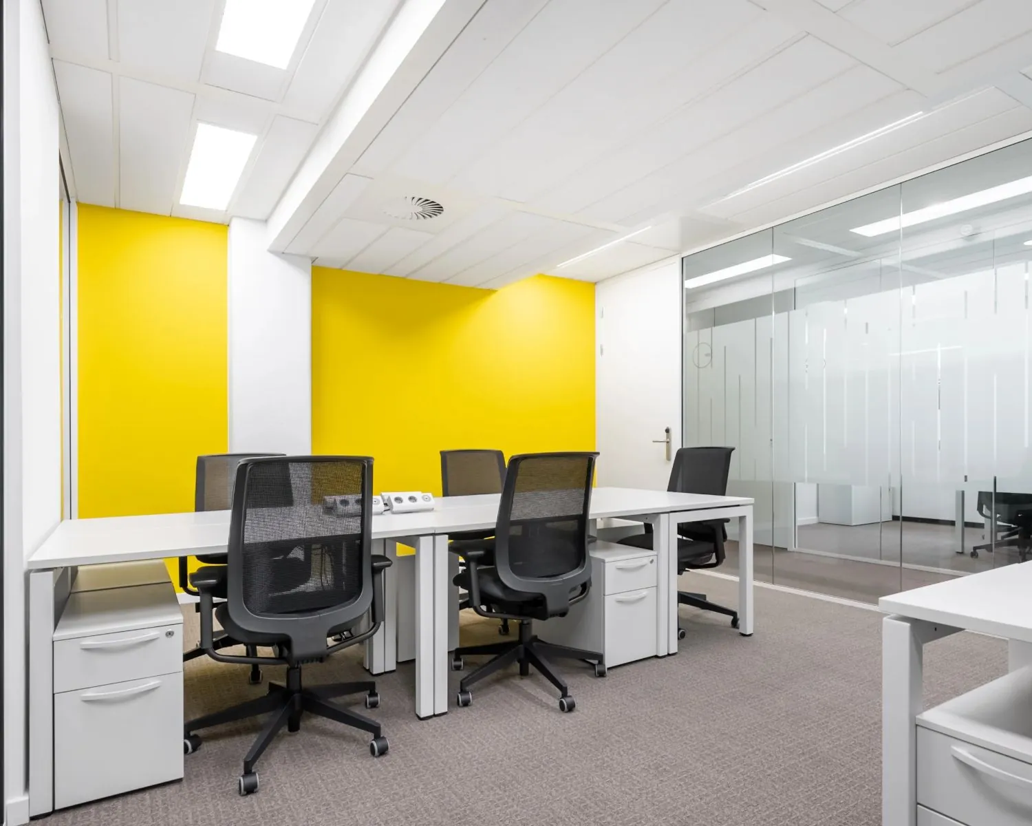 Moderne kantoorruimte met vier bureaus, zwarte bureaustoelen en een gele accentmuur.