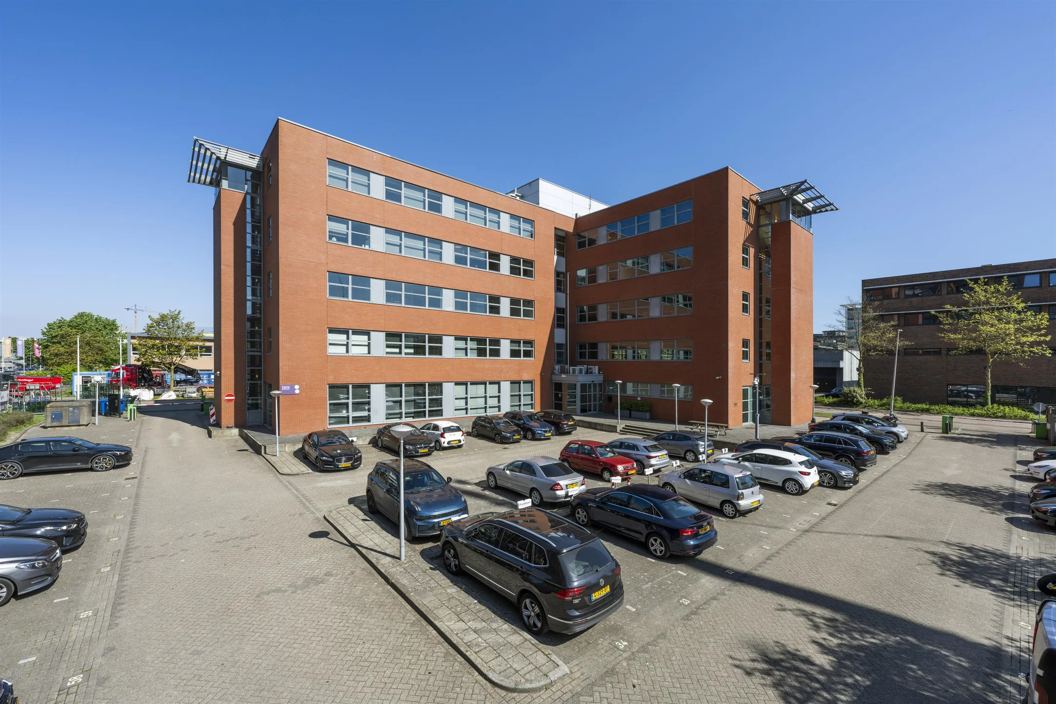 Modern red-brick office building labeled "549_2160" with a surrounding parking lot full of cars under a clear blue sky.