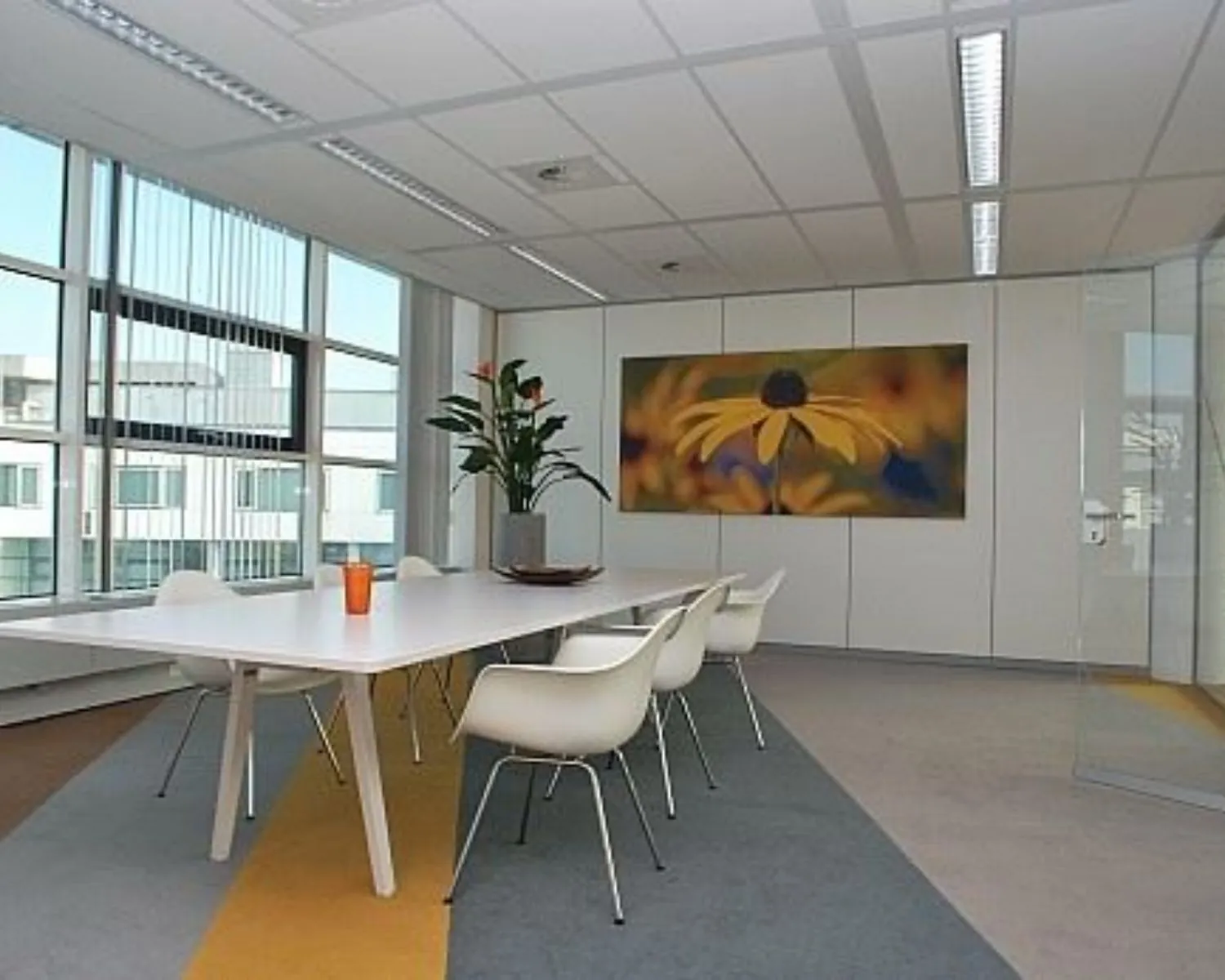 Moderne vergaderruimte met een lange witte tafel, witte stoelen, een grote plant en een kleurrijk bloemenkunstwerk aan de muur.