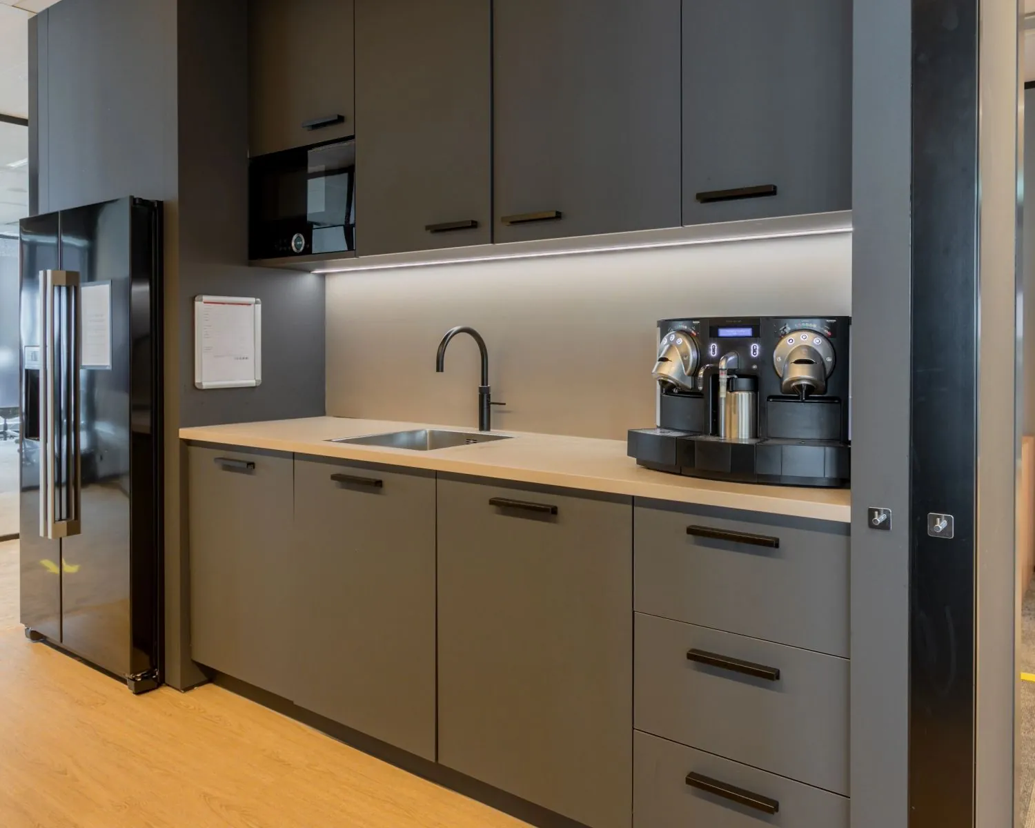 Moderne kantoorpantry met zwarte kasten, ingebouwde apparatuur en een dubbele koffiemachine.