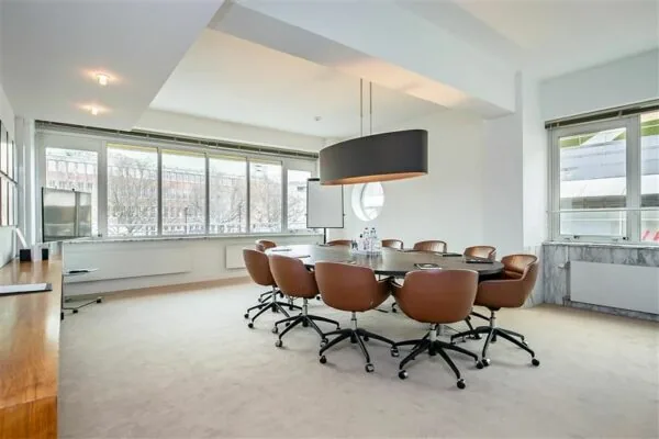 Moderne vergaderruimte met een ovale tafel, bruine stoelen en grote ramen met uitzicht op een kantoorgebouw.