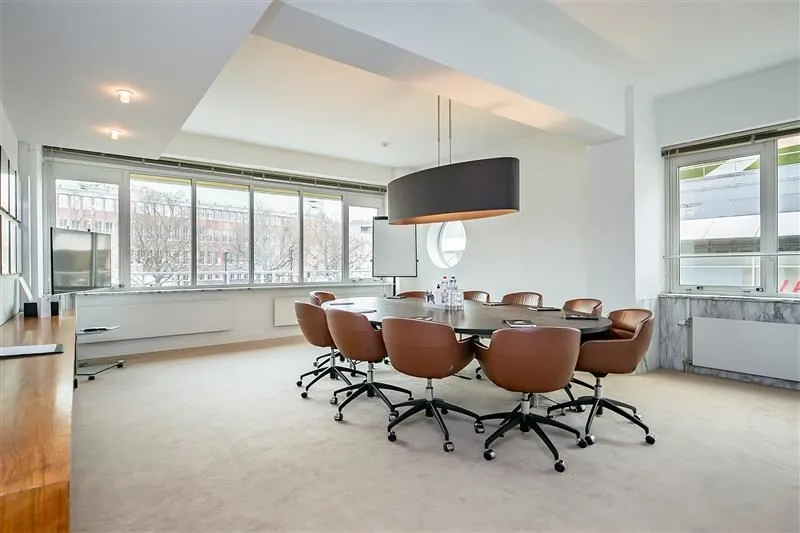 Moderne vergaderruimte met een ovale tafel, bruine stoelen en grote ramen met uitzicht op een kantoorgebouw.