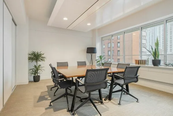 Moderne vergaderruimte met een houten tafel, acht zwarte stoelen, grote ramen en groene kamerplanten.