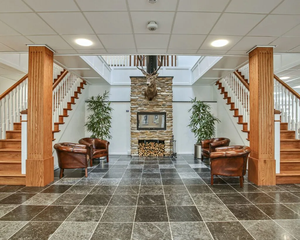 Ruime ontvangstruimte met een open haard, leren fauteuils, houten trap en een opgezette hertenkop aan de muur.
