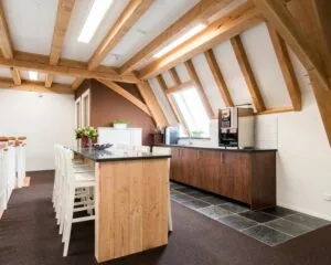 Moderne kitchenette met houten balkenplafond, lange tafel met stoelen en koffiemachine onder een dakraam.