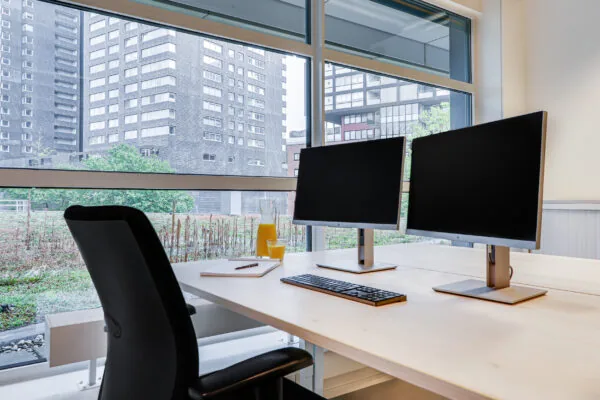 Lichte coworking space met twee computerschermen, een toetsenbord en een glazen kan sinaasappelsap bij het raam.