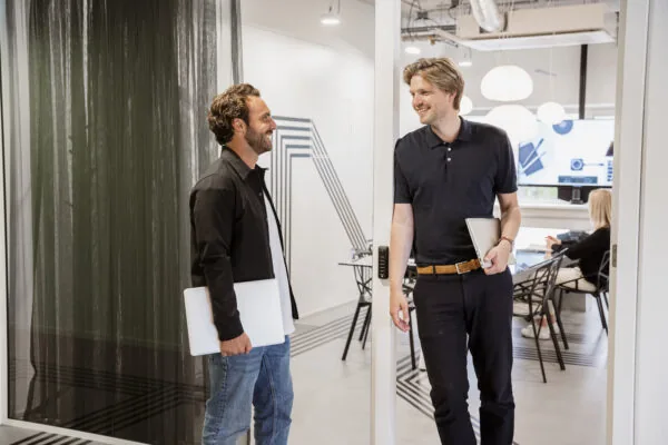 Twee mannen met laptops praten en lachen in een modern kantoor van HNK Amsterdam Schinkel.