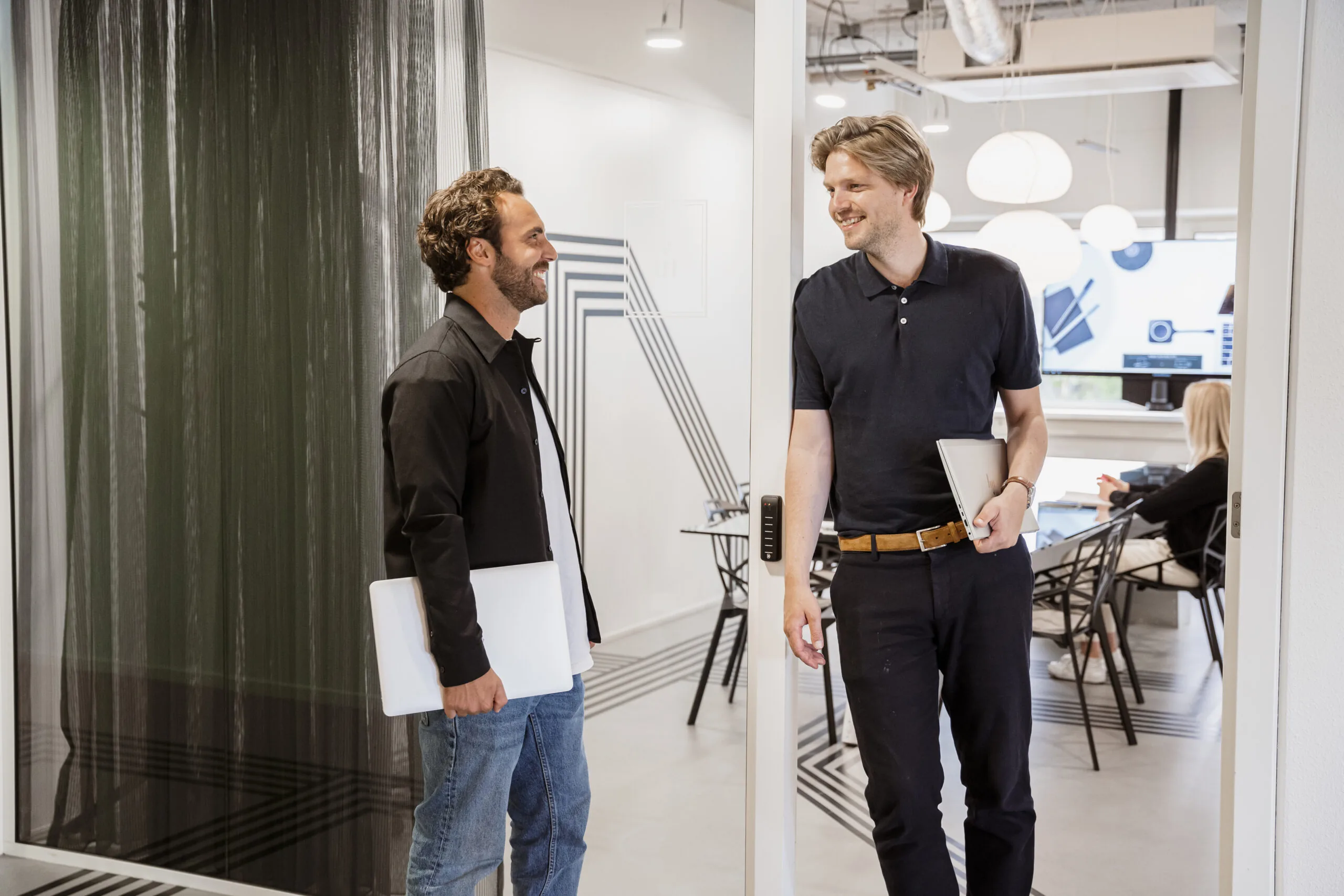 Twee mannen met laptops praten en lachen in een modern kantoor van HNK Amsterdam Schinkel.