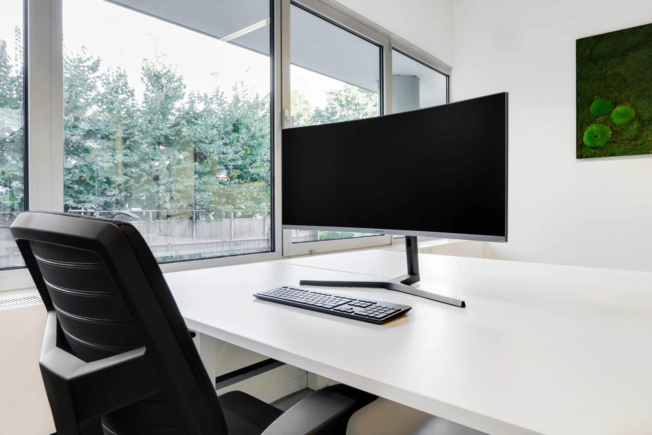 Een moderne co-working werkplek in Utrecht West met een groot beeldscherm, toetsenbord en bureaustoel bij een raam met uitzicht op groen.