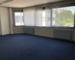 Lege kantoorruimte met blauwe vloerbedekking en grote ramen met verticale lamellen aan de Hooilaan 1 in Breda.