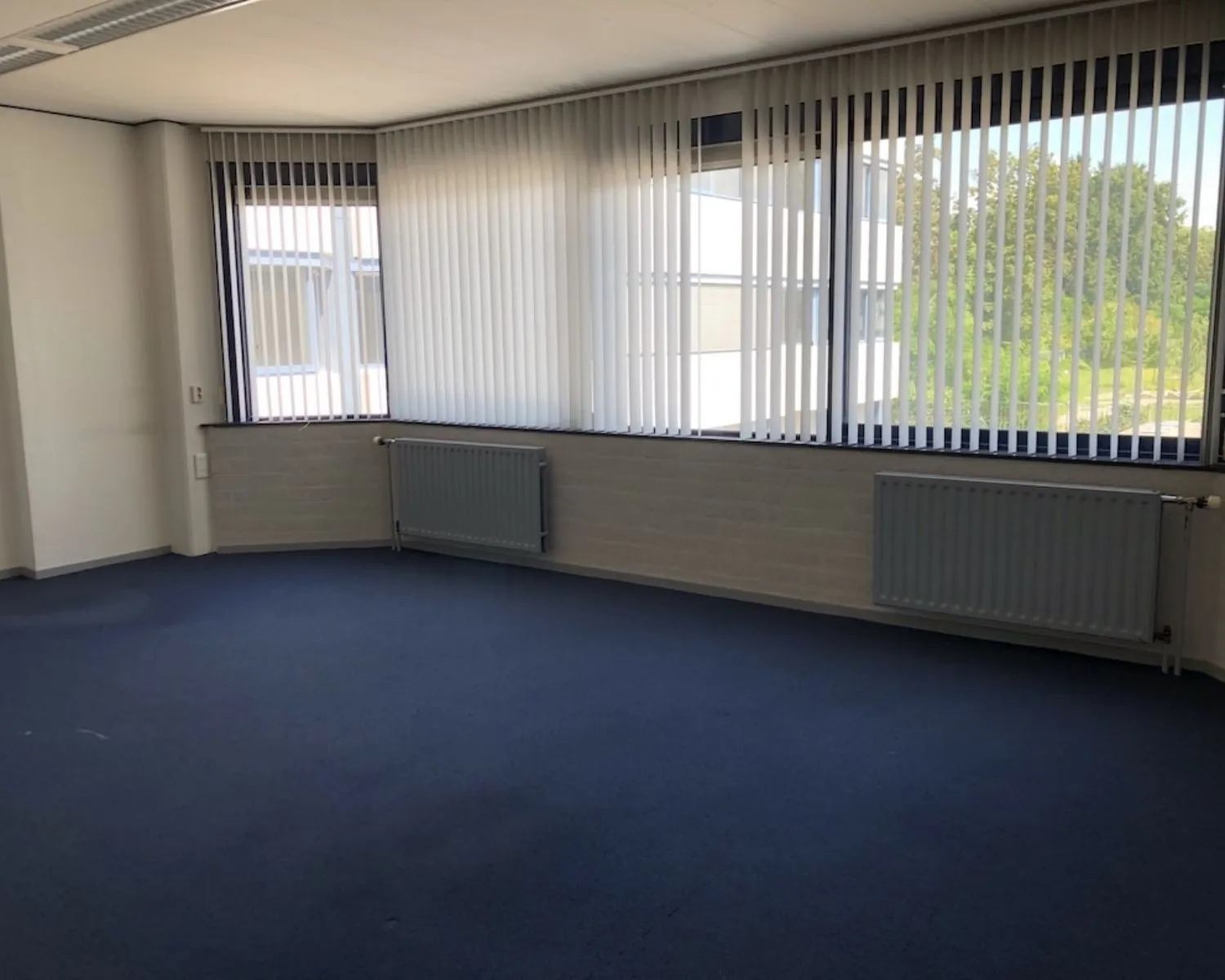 Lege kantoorruimte met blauwe vloerbedekking en grote ramen met verticale lamellen aan de Hooilaan 1 in Breda.