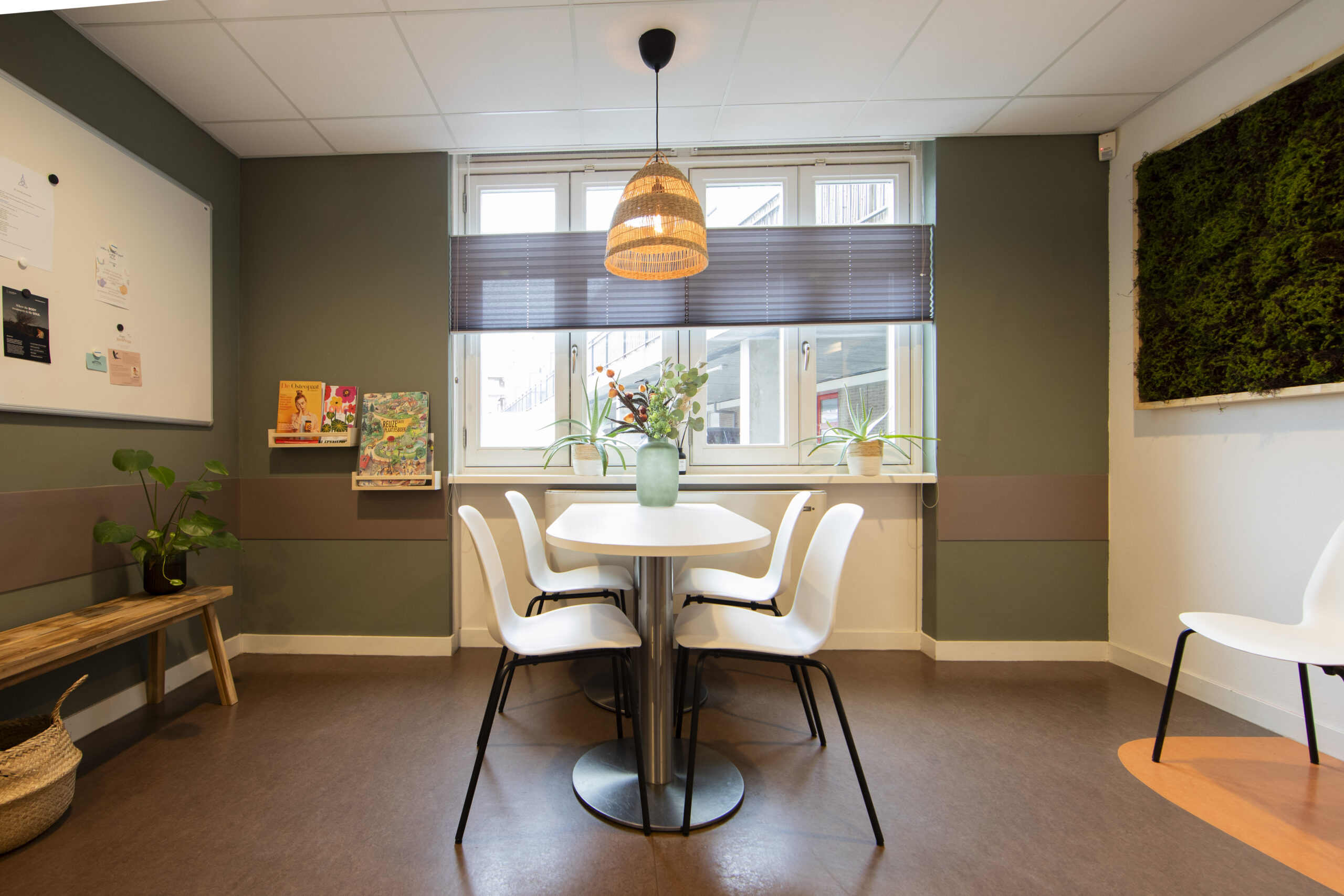 Een moderne wachtruimte met witte stoelen rondom een tafel, decoratieve planten en een hanglamp.