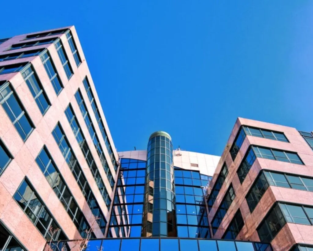 Vooraanzicht van een modern kantoorgebouw met glazen gevel en roze stenen elementen tegen een blauwe lucht.