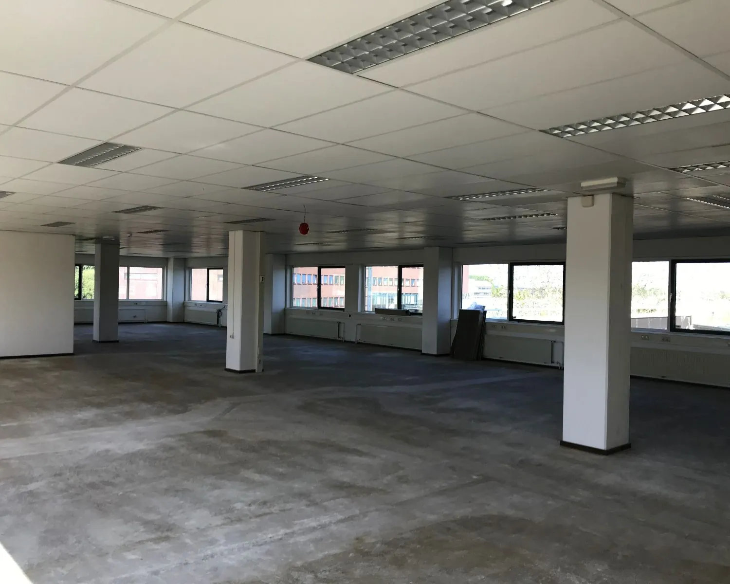 Lege kantoorruimte met systeemplafond, betonnen vloer en grote ramen rondom.