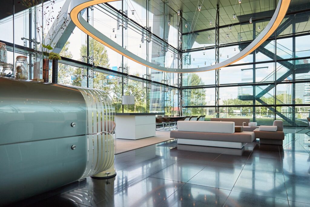 Moderne lobby met glazen wanden, designmeubilair en een opvallend verlicht plafond aan de Amstelveenseweg.