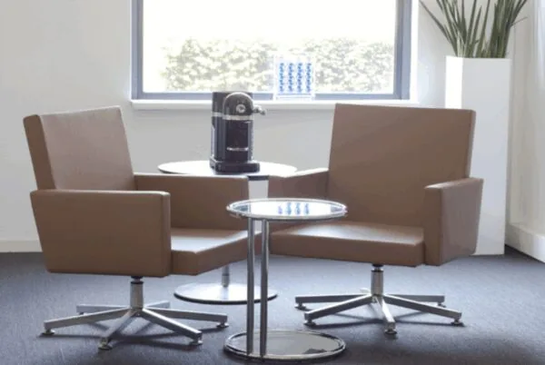 Twee moderne bruine stoelen met bijzettafels en een koffiemachine in een lichte wachtruimte aan de Arlandaweg.