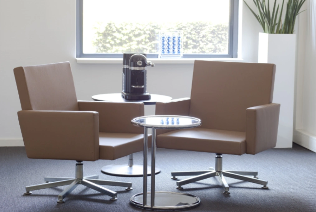 Twee moderne bruine stoelen met bijzettafels en een koffiemachine in een lichte wachtruimte aan de Arlandaweg.