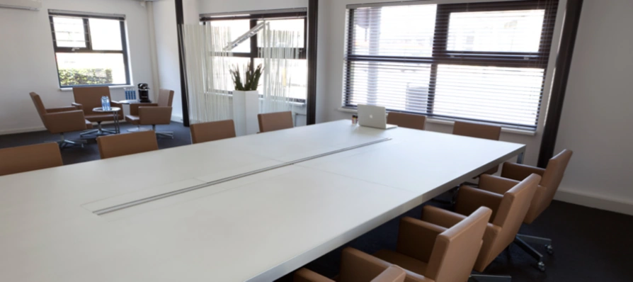 Vergaderruimte aan de Arlandaweg met een lange witte tafel, bruine stoelen en grote ramen met jaloezieën.