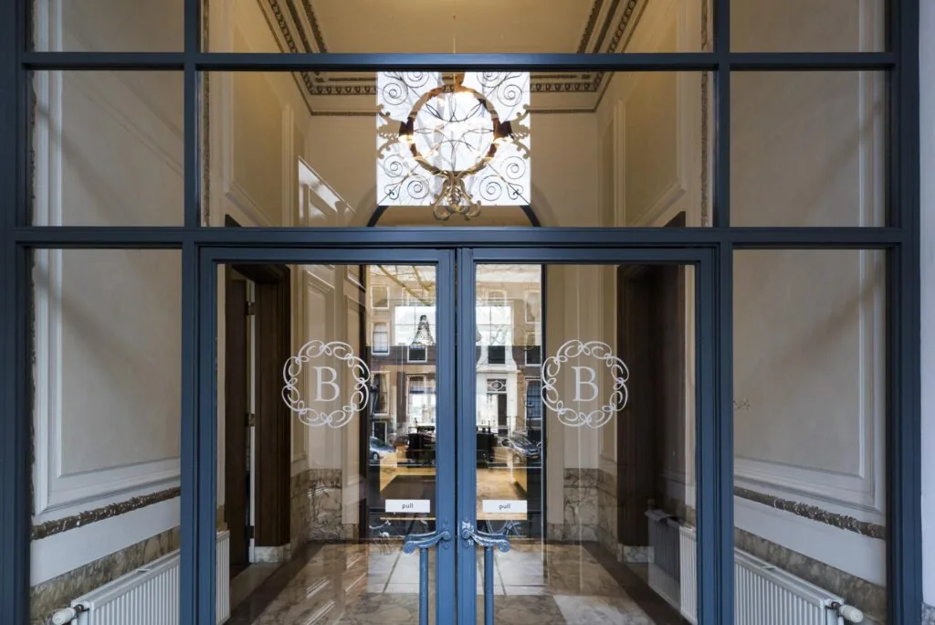 Glazen ingang met dubbele deuren en het embleem "B" aan de Herengracht, met zicht op een stijlvolle binnenruimte.