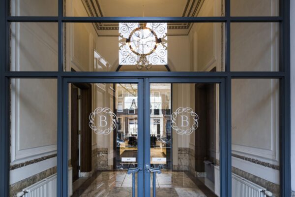 Glazen ingang met dubbele deuren en het embleem "B" aan de Herengracht, met zicht op een stijlvolle binnenruimte.