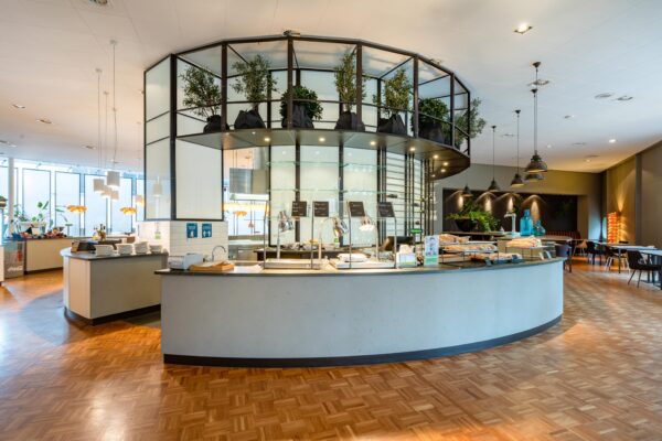 Een moderne en lichte cafetaria met een centrale toonbank en groene planten.