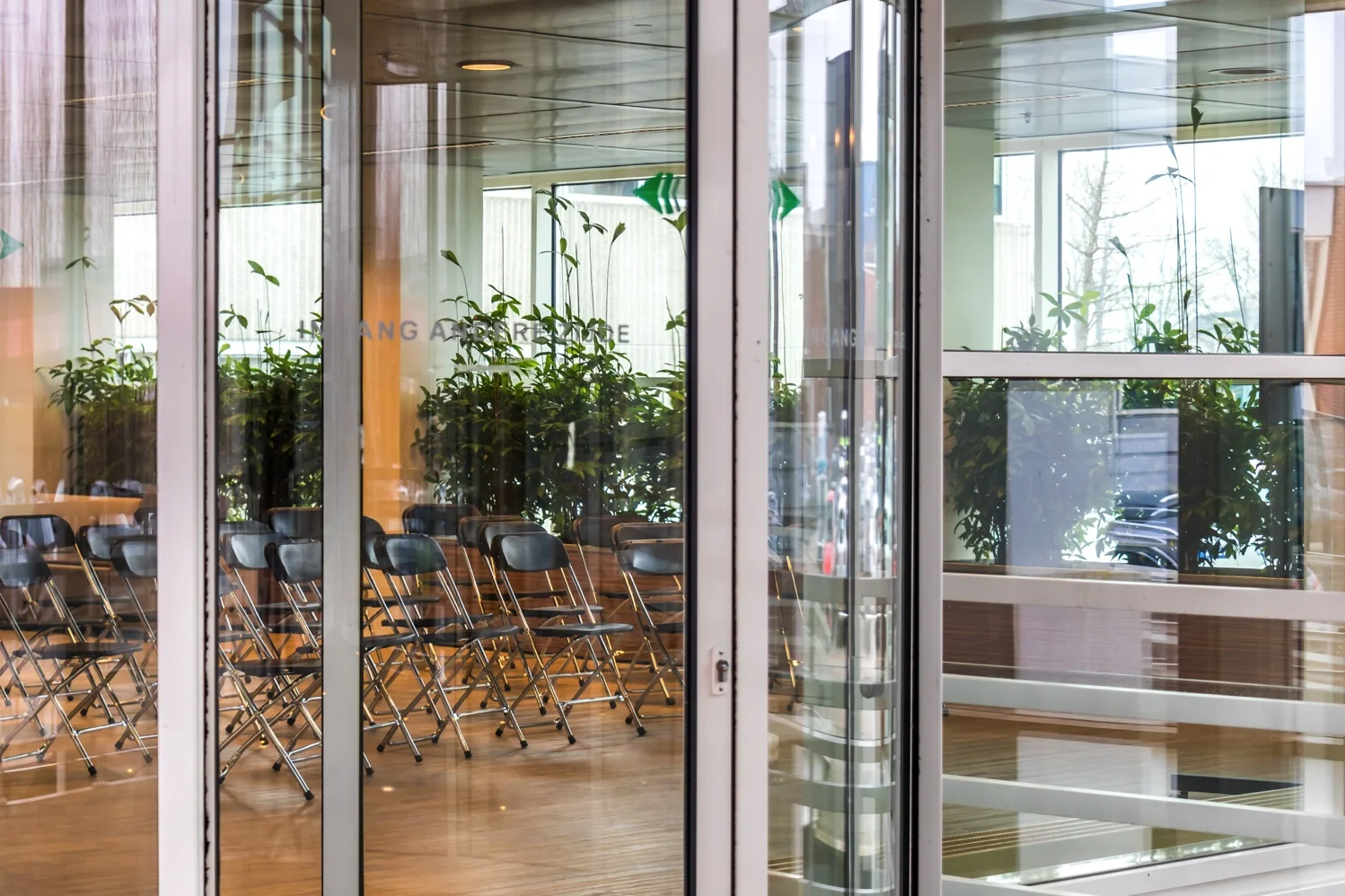 Ingang van een modern gebouw aan de Reactorweg met zicht op een wachtruimte met opklapstoelen en groene planten.