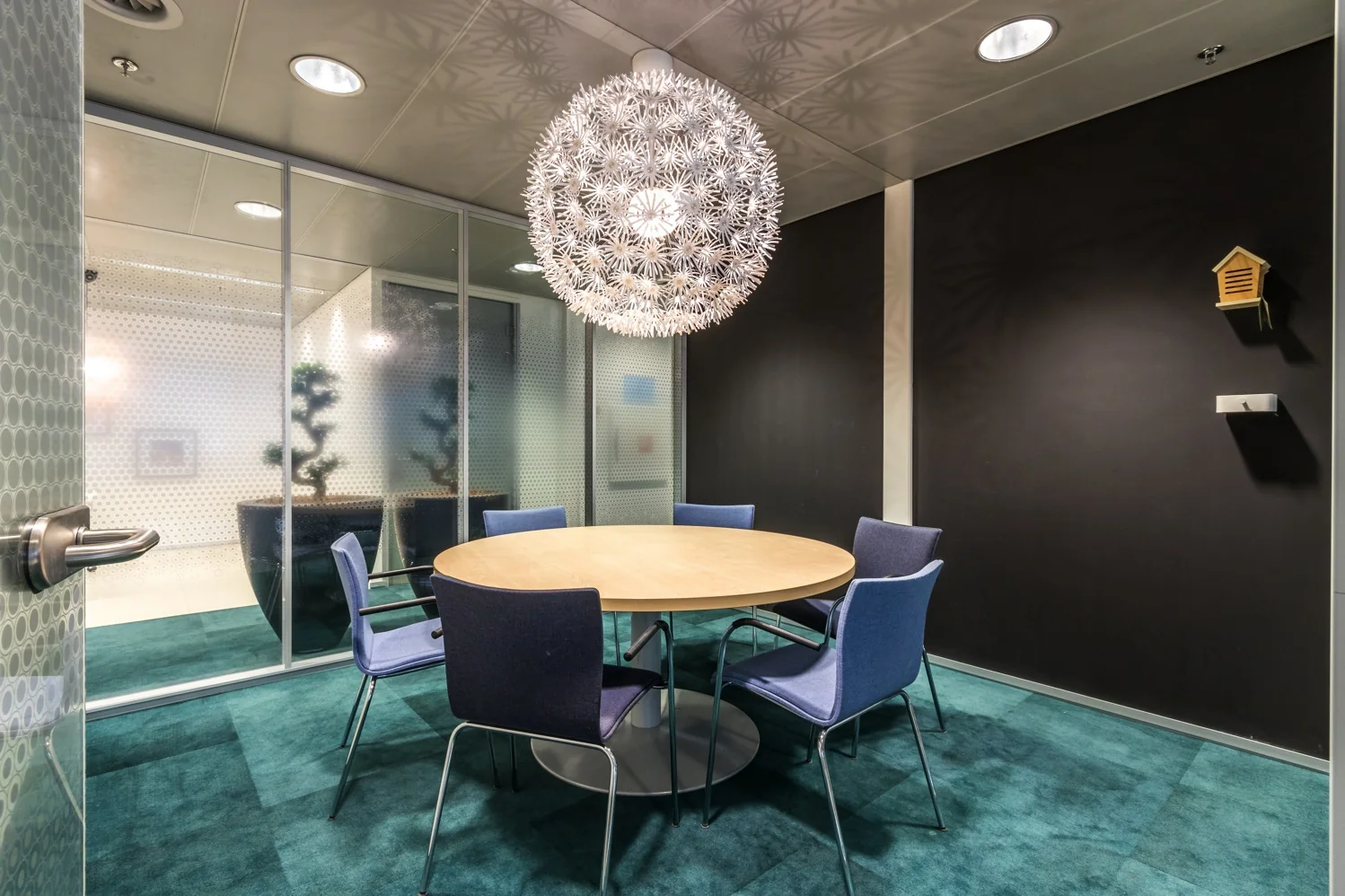 Vergaderruimte met een ronde houten tafel, zes blauwe stoelen en een opvallende bolvormige hanglamp aan het plafond.