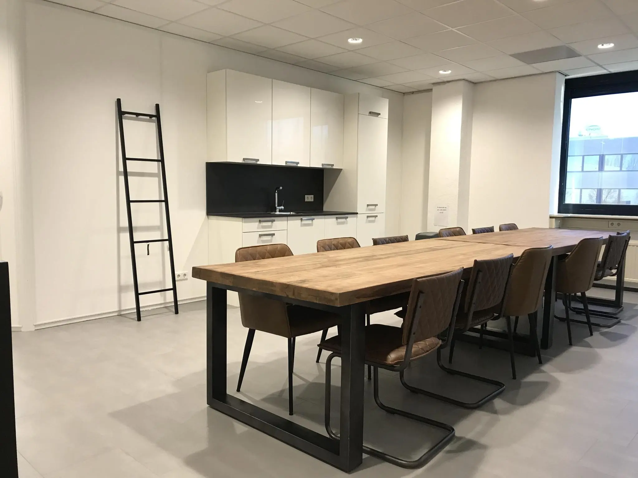 Vergaderruimte met lange houten tafel, leren stoelen, kitchenette en een zwarte ladder tegen de muur.