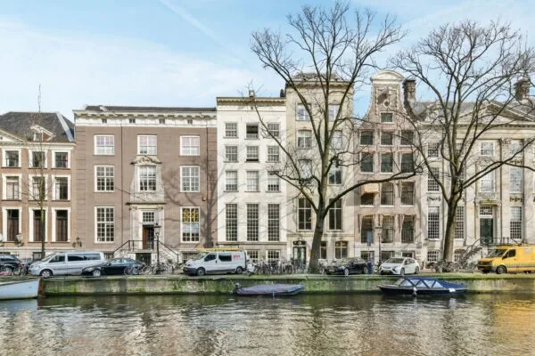 Gevels van historische grachtenpanden aan de Herengracht in Amsterdam met geparkeerde auto's en boten langs het water.