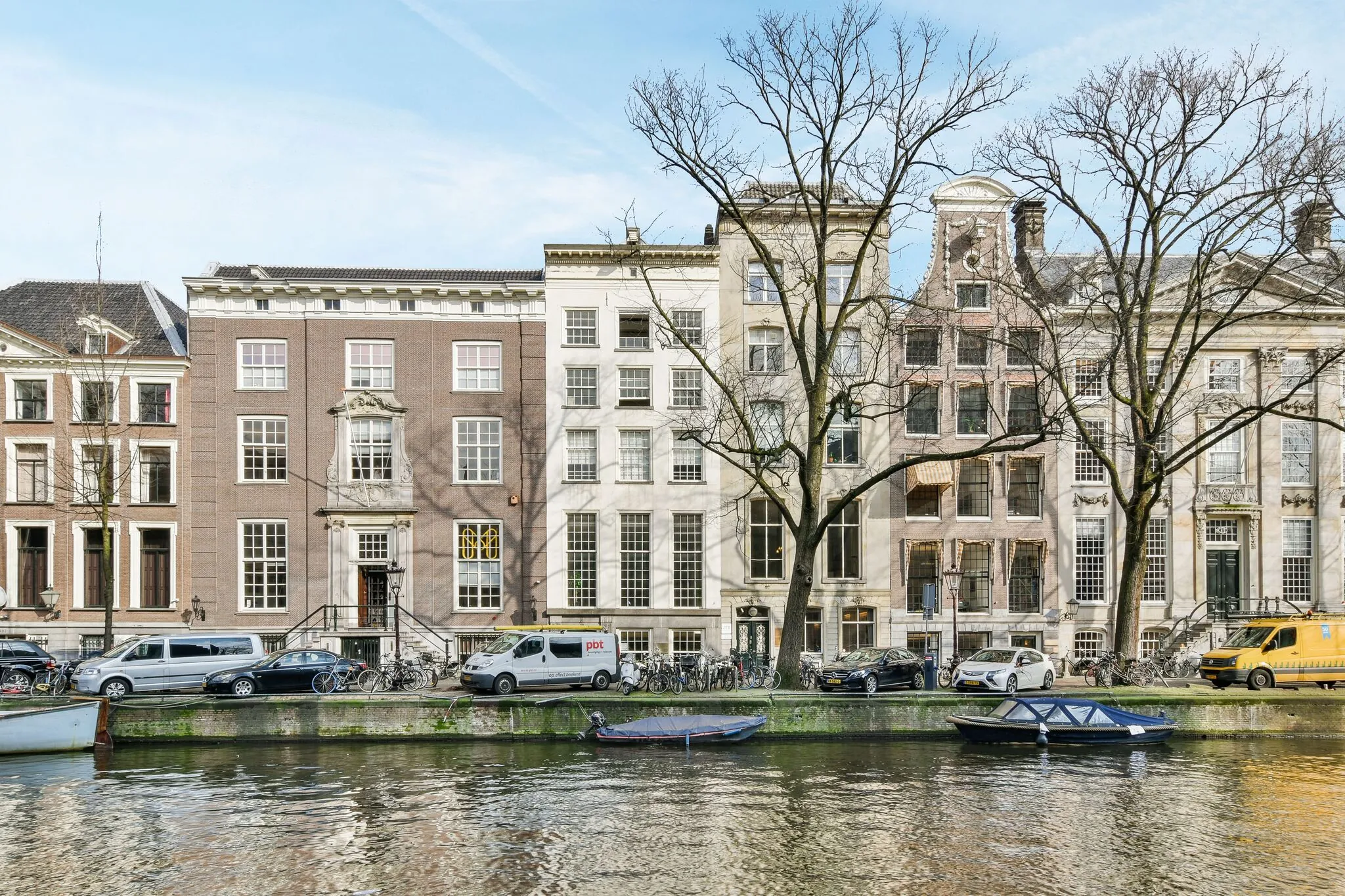 Gevels van historische grachtenpanden aan de Herengracht in Amsterdam met geparkeerde auto's en boten langs het water.