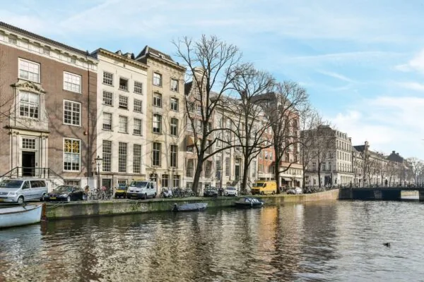Zicht op historische panden aan de Herengracht in Amsterdam met geparkeerde auto's, fietsen en boten langs de gracht.